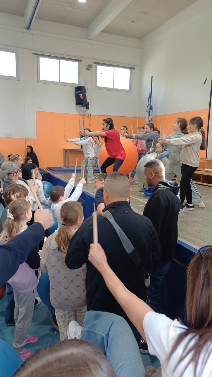 הילה מובילה תיפוף מול כדור כתום ענק, וילדים והורים מתופפים איתה