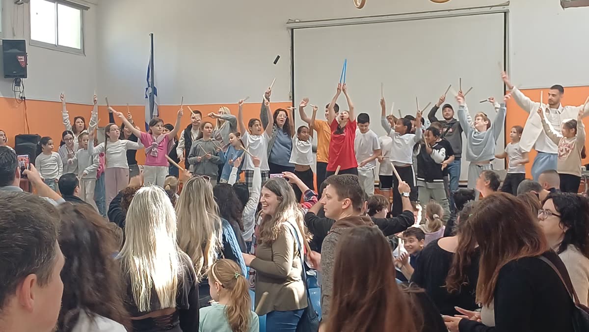 ילדים ומבוגרים מרימים מקלות תיפוף בשמחה, והילה במרכז